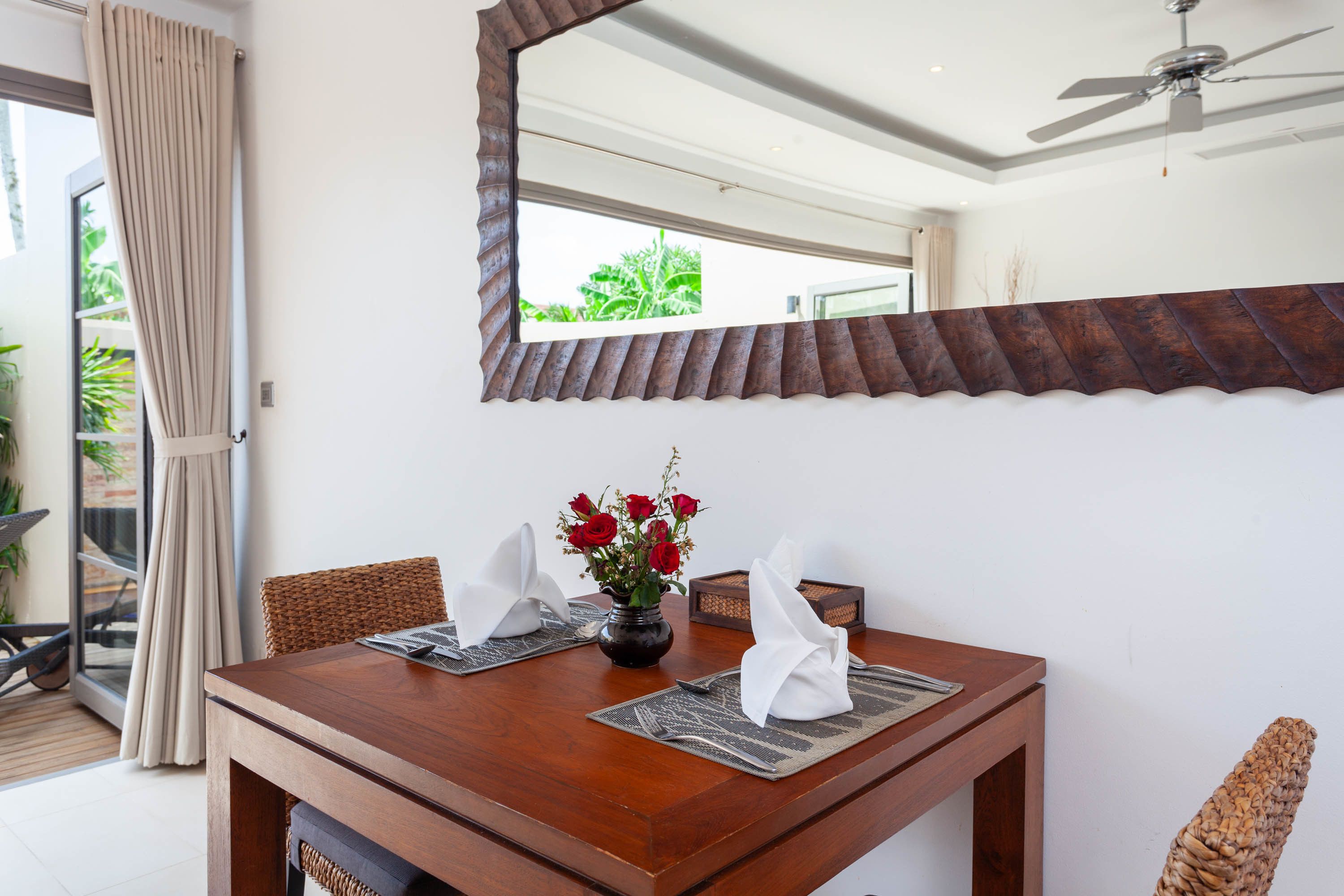 A dining table with placemats, a vase with red flowers, and a decorative mirror above it.