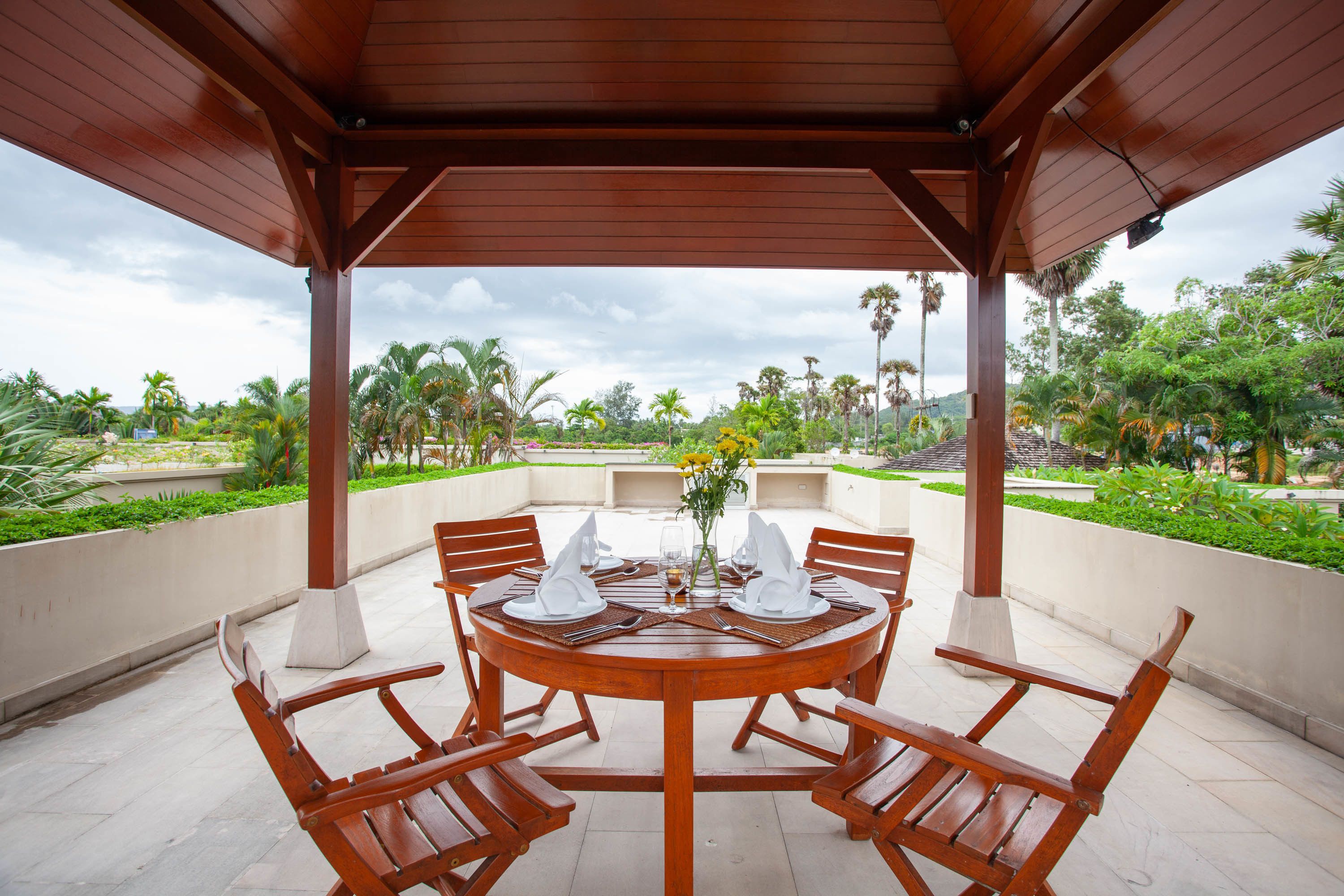 A wooden outdoor dining area with a round table set for four, surrounded by chairs, under a wooden pergola.