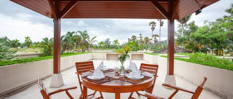 A wooden outdoor dining area with a round table set for four, surrounded by chairs, under a wooden pergola.