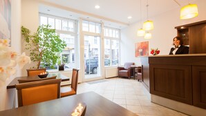 En hotellobby med en receptionsdisk i trä, en receptionist och ett stort fönster med utsikt över gatan. Det finns krukväxter och en matplats med ett bord och stolar.