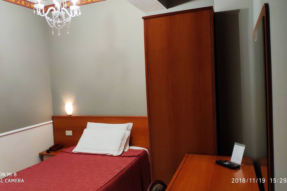 Una habitación de hotel con cama, mesita de noche y escritorio con silla. Hay una araña en el techo y una lámpara fijada a la pared.