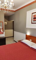 Habitación de hotel con cama, mesita de noche, espejo, candelabro, un cuadro en la pared y puerta que da a un baño.
