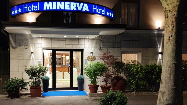 Exterior do Hotel Minerva, com uma placa azul, entrada dupla e vasos de plantas na frente.