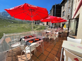 Una terraza al aire libre con sombrillas rojas, sillas y mesas blancas, con vista a un paisaje montañoso.