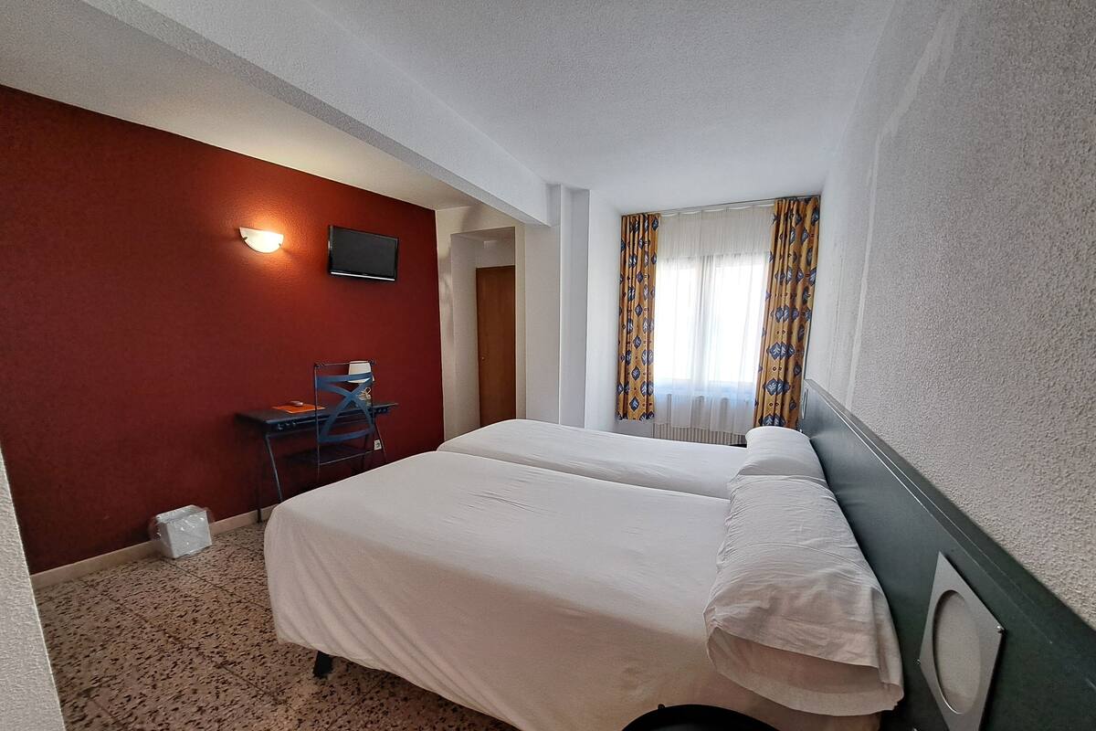 Habitación de hotel con cama, escritorio, silla, televisor y ventana con cortinas.