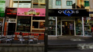 Ein Hotel mit mehreren Etagen, Schilder mit den Aufschriften 'CHAMBRES' und 'HABITATIONS' zur Kennzeichnung verfügbarer Zimmer, sowie eine Schaufensterfront mit den Schildern 'BUFFET' und 'LIURE', die Speiseoptionen andeuten.