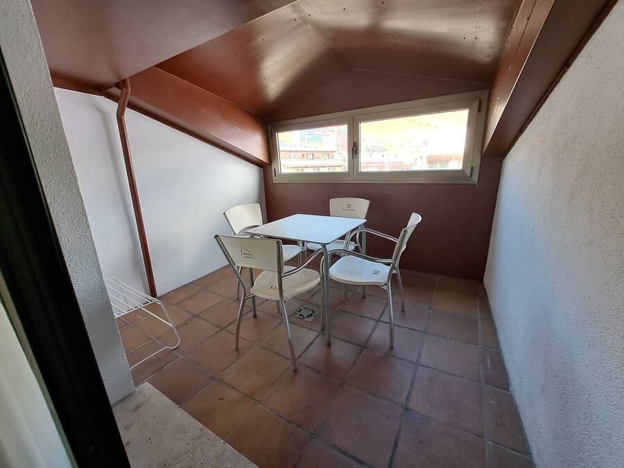Uma varanda pequena e fechada, com mesa e cadeiras, uma janela com vista para edifícios e um chão de azulejos.