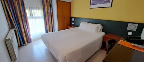 Habitación de hotel con cama, escritorio, silla, radiador, cortinas, puerta y un cuadro en la pared.