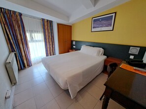 Hotelkamer met een bed, bureau, stoel, radiator, gordijnen, deur en een lijst met een afbeelding aan de muur.