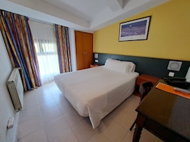 Habitación de hotel con cama, escritorio, silla, radiador, cortinas, puerta y un cuadro en la pared.