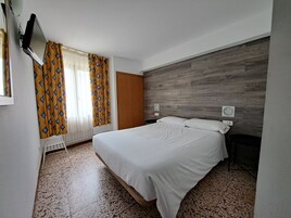 A hotel room with a bed, a wooden accent wall, a TV mounted on the wall, and a window with curtains.