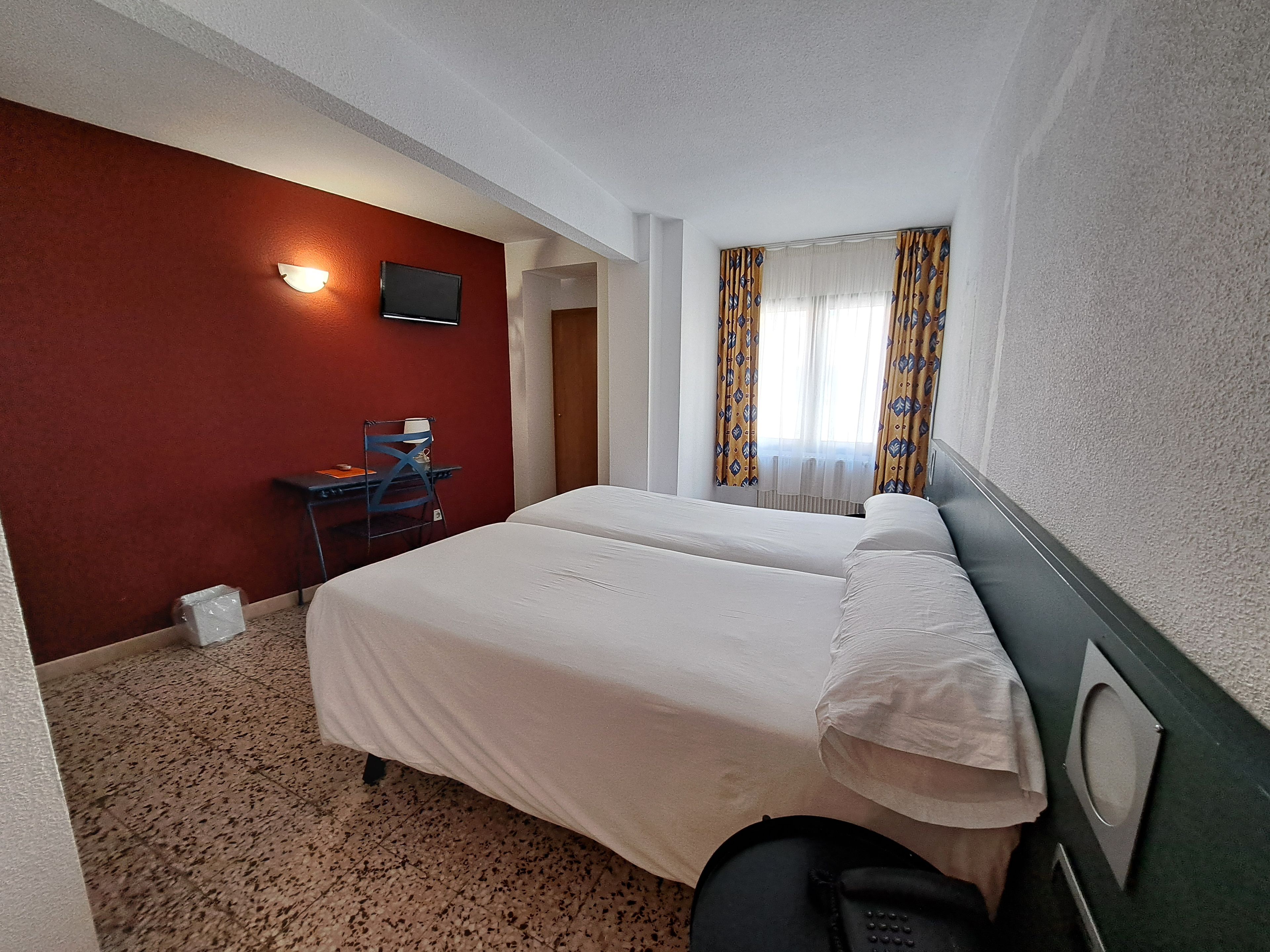 A hotel room with a bed, a desk, a chair, a TV, and a window with curtains.