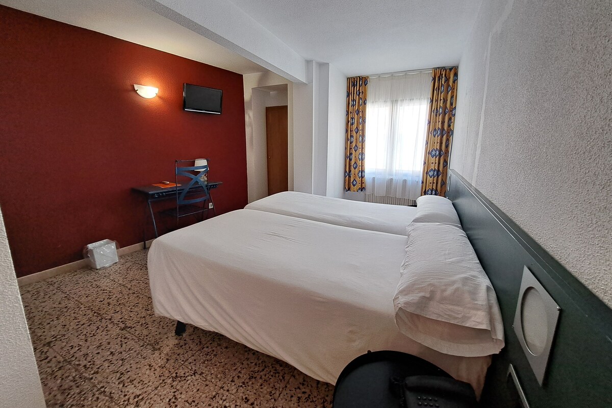 A hotel room with a bed, a desk, a chair, a TV, and a window with curtains.