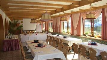 A dining area with long tables and chairs, wooden beams on the ceiling, and large windows with curtains.