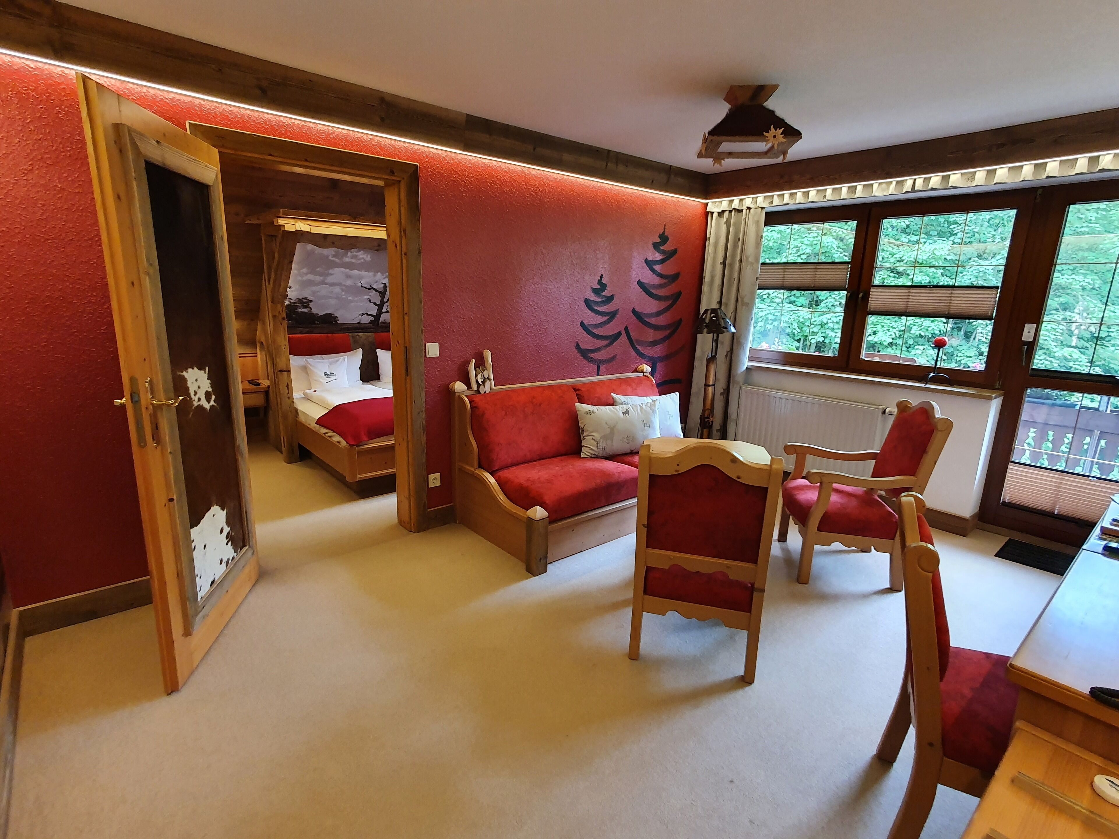 A room with a bed, a red sofa, two wooden chairs, and a window with a view of greenery.