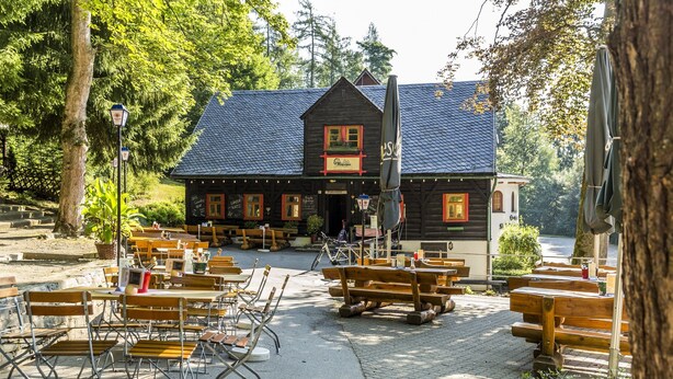 A rustic wooden building with outdoor seating and a large umbrella. - Hotel und Restaurant Köhlerhütte (Gruenhain-Beierfeld)