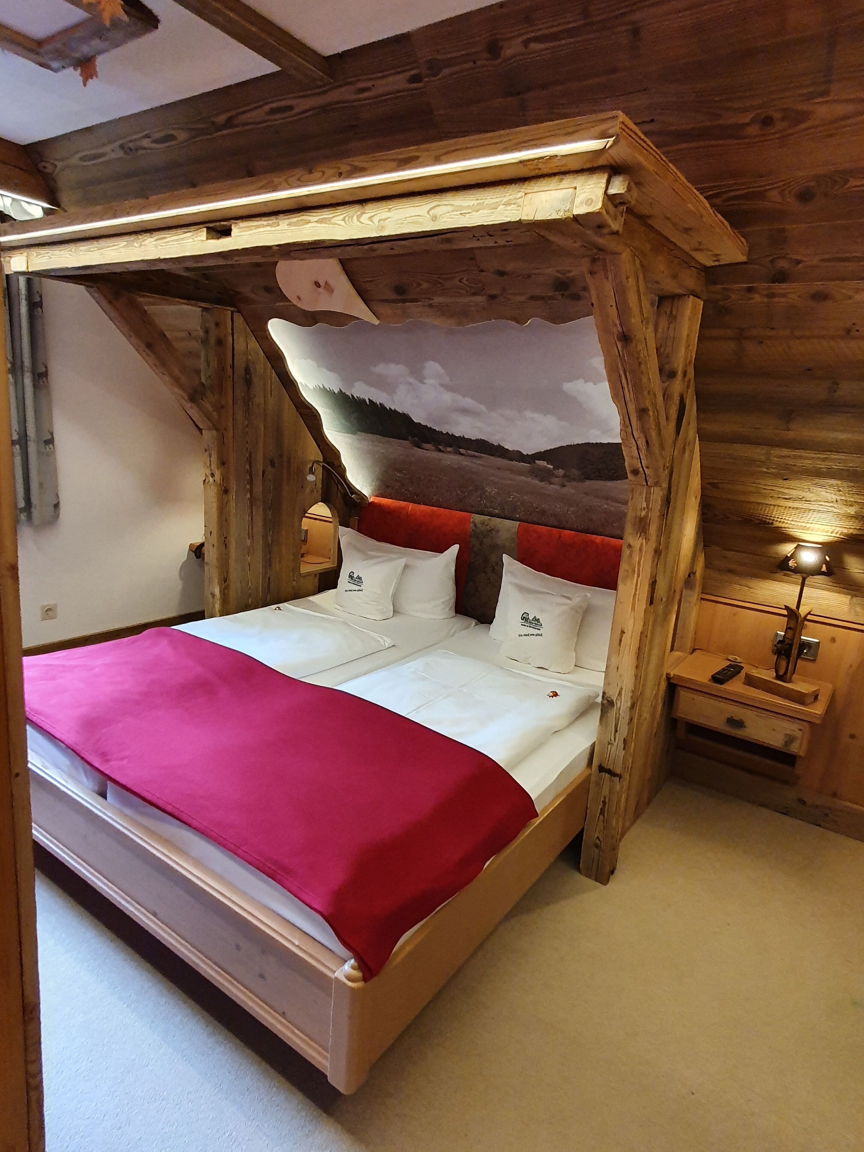 Una cama con dosel de madera con una manta roja, almohadas blancas y un mural de paisaje en la pared de fondo.