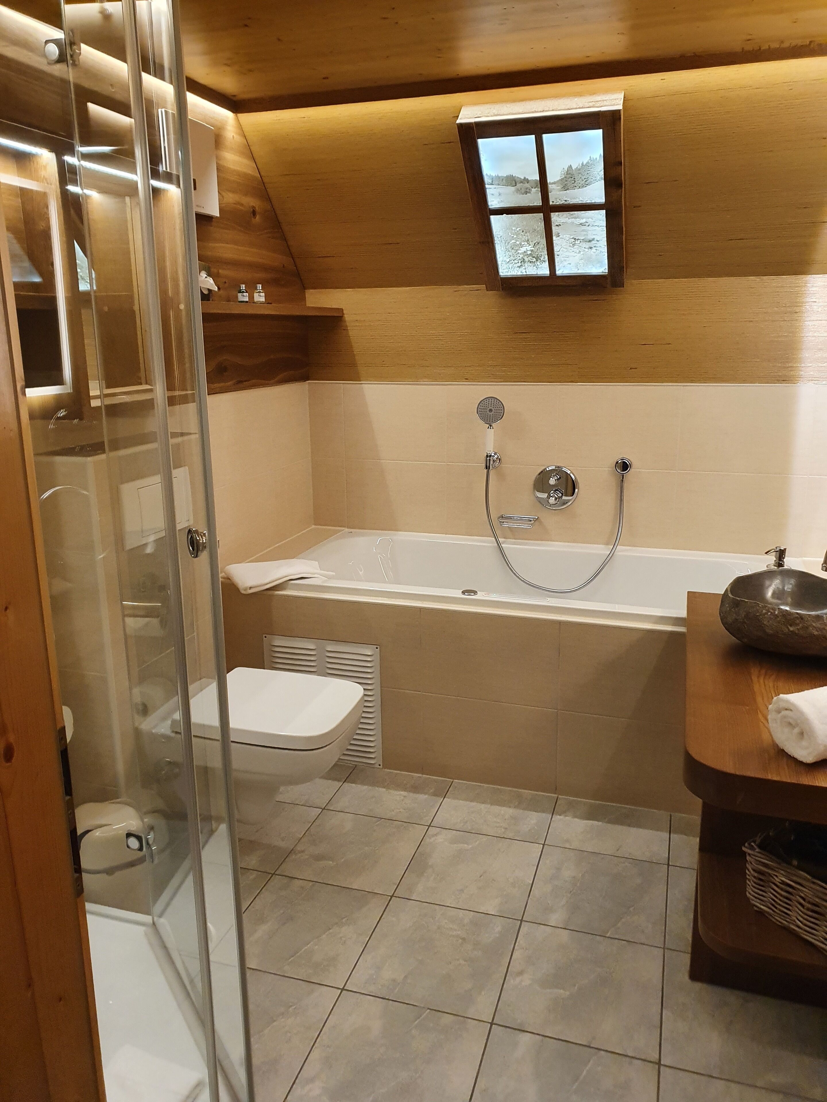 Un baño moderno con ducha envuelta en cristal, una bañera independiente y un tocador de madera con lavabo de piedra.