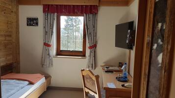 Una habitación con una cama, un escritorio, una silla, un ventanal con cortinas, un televisor y un pequeño cuadro en la pared.