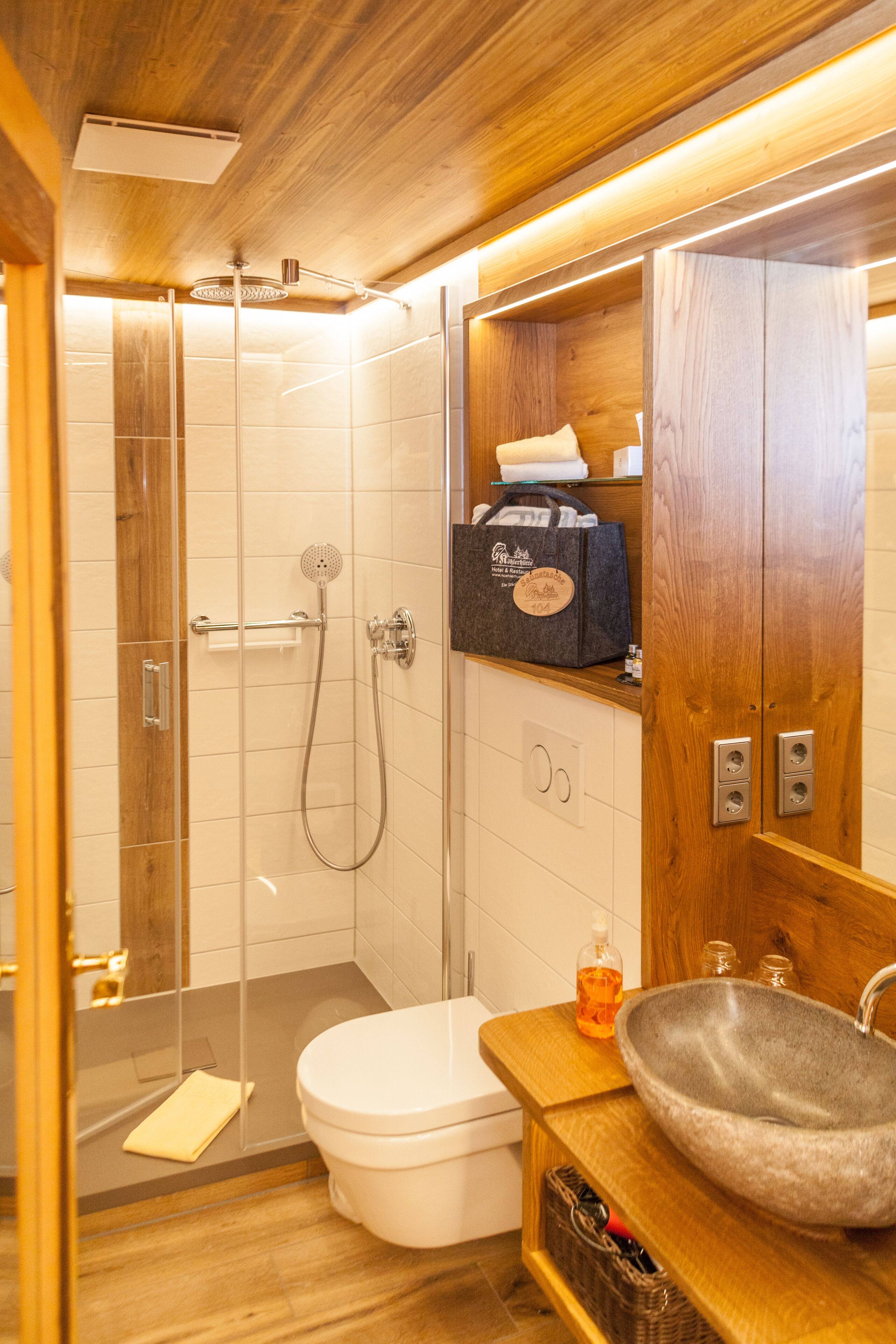 Een compacte badkamer met een douchecabine van glas, een wit toilet en een houten wastafel met een stenen wasbak.