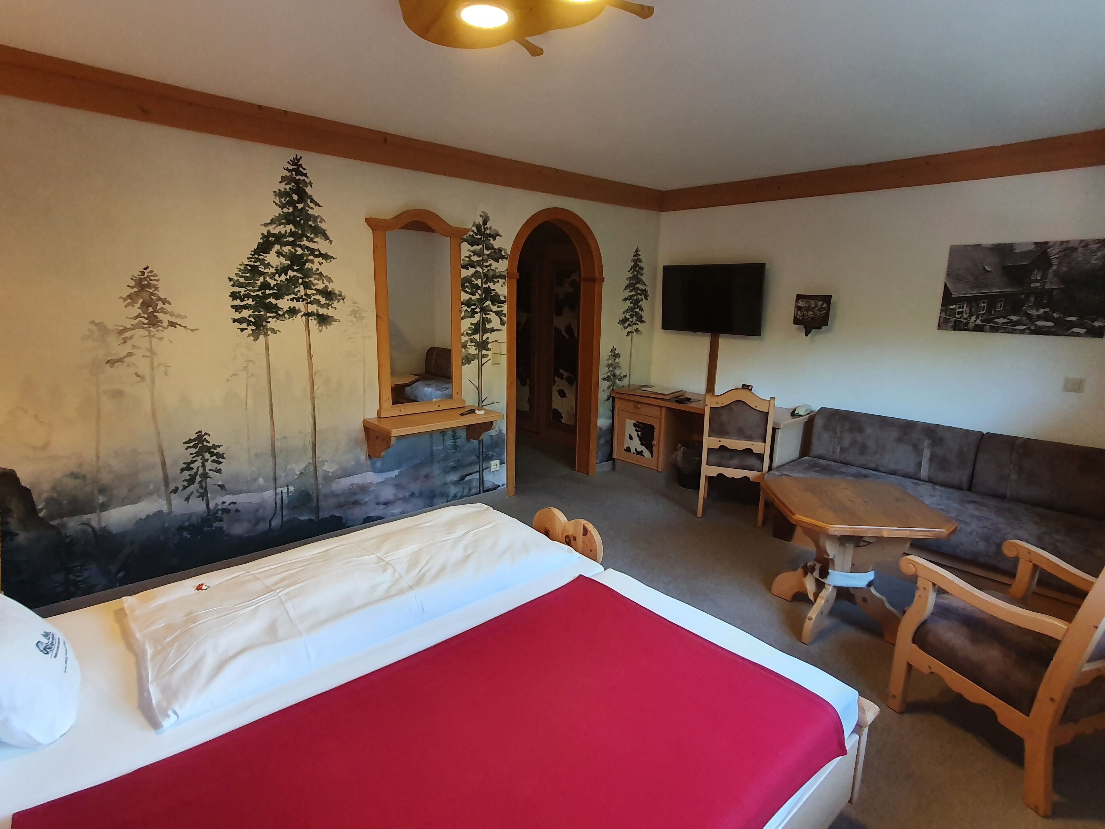 A hotel room with a bed, a red blanket, a wooden desk, a TV, a sofa, and a mural of trees.