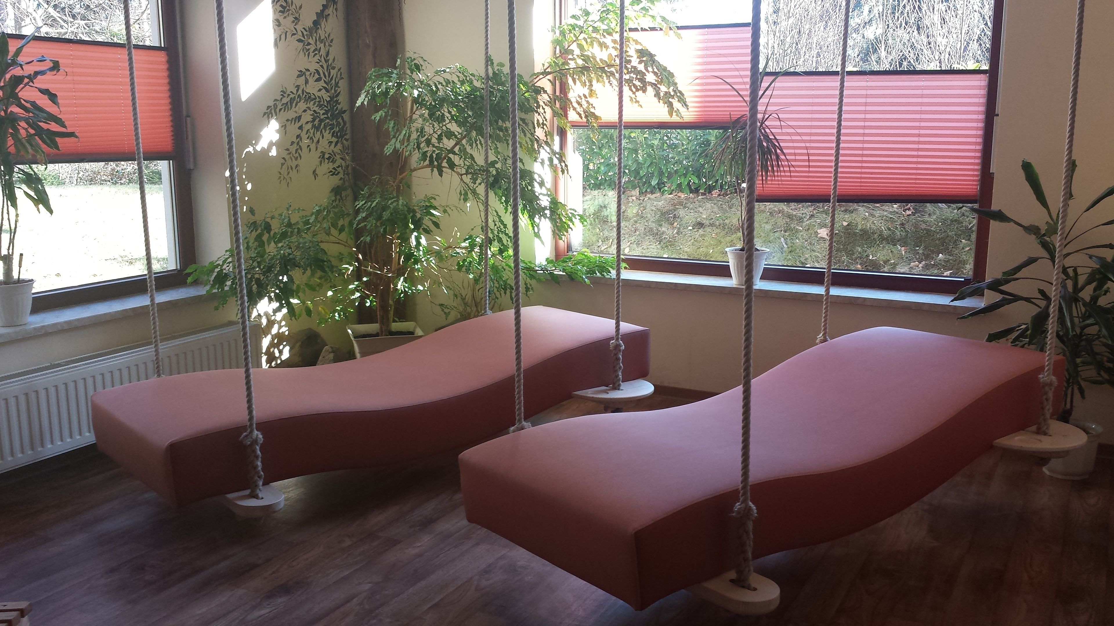 A room with two red cushioned swings, a potted plant, and a view of the outside through a window with a red blind.