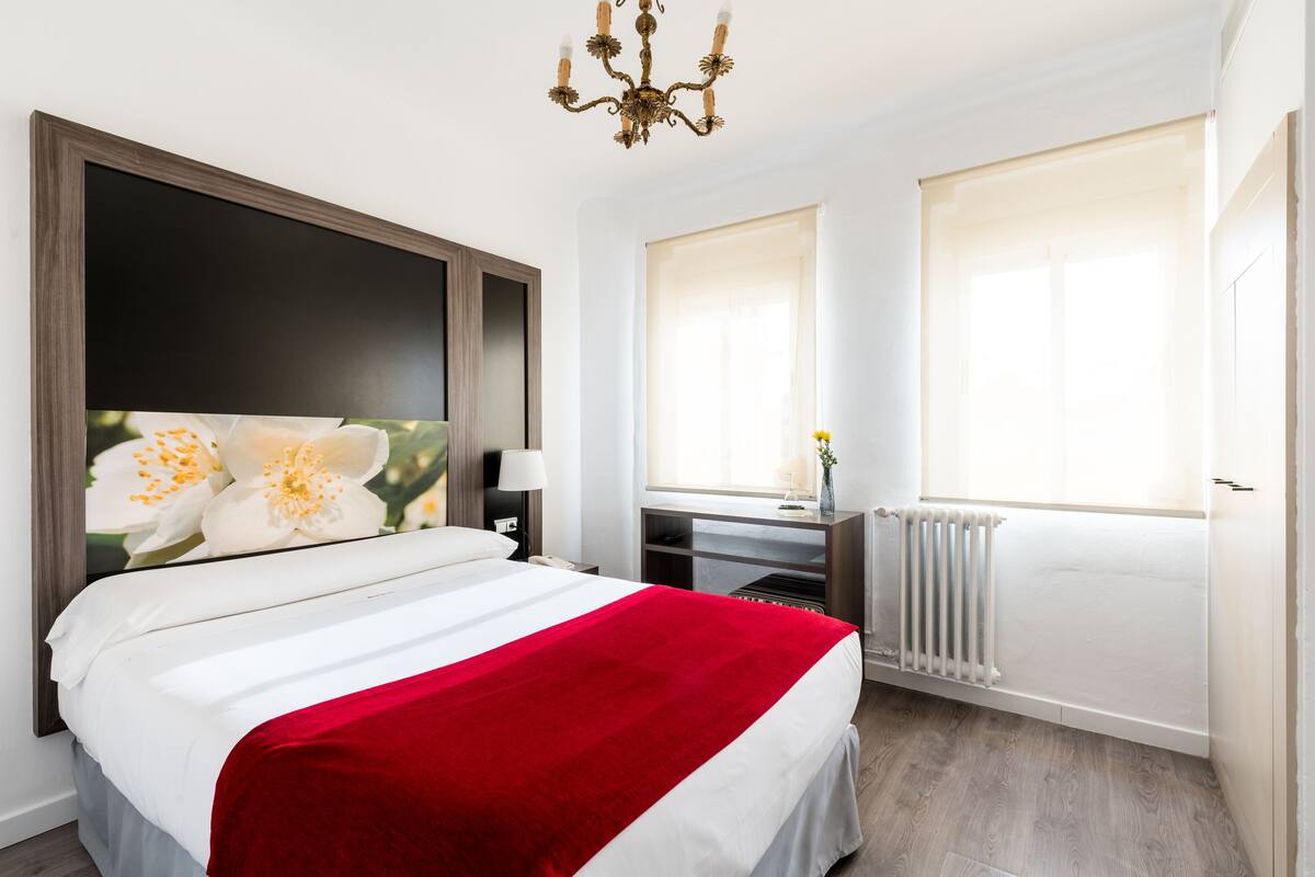 Ein Schlafzimmer mit einem Bett, einem Nachttisch, einer Kommode, einem Spiegel und einem Fenster mit Jalousien.