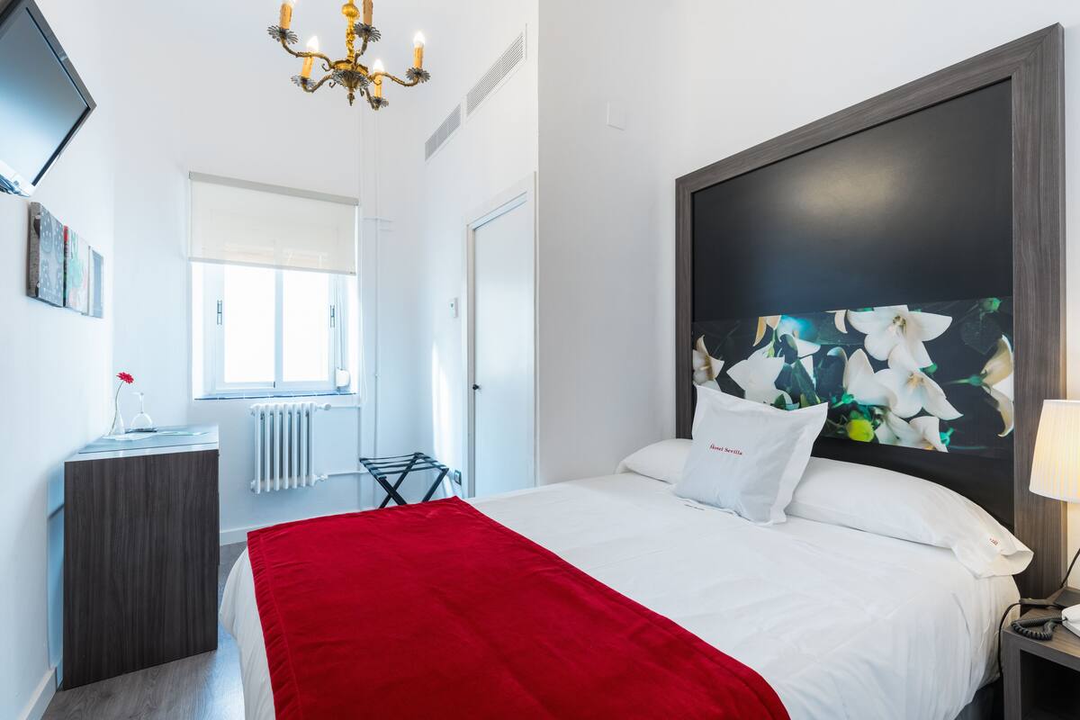 Ein Hotelzimmer mit einem Bett, einer roten Tagesdecke, einem großen Blumenbild an der Wand, einem Nachttisch mit Lampe und einem Fenster mit Jalousien.