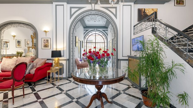 A grand lobby with a red sofa, a glass table with flowers, a staircase, and a chandelier.