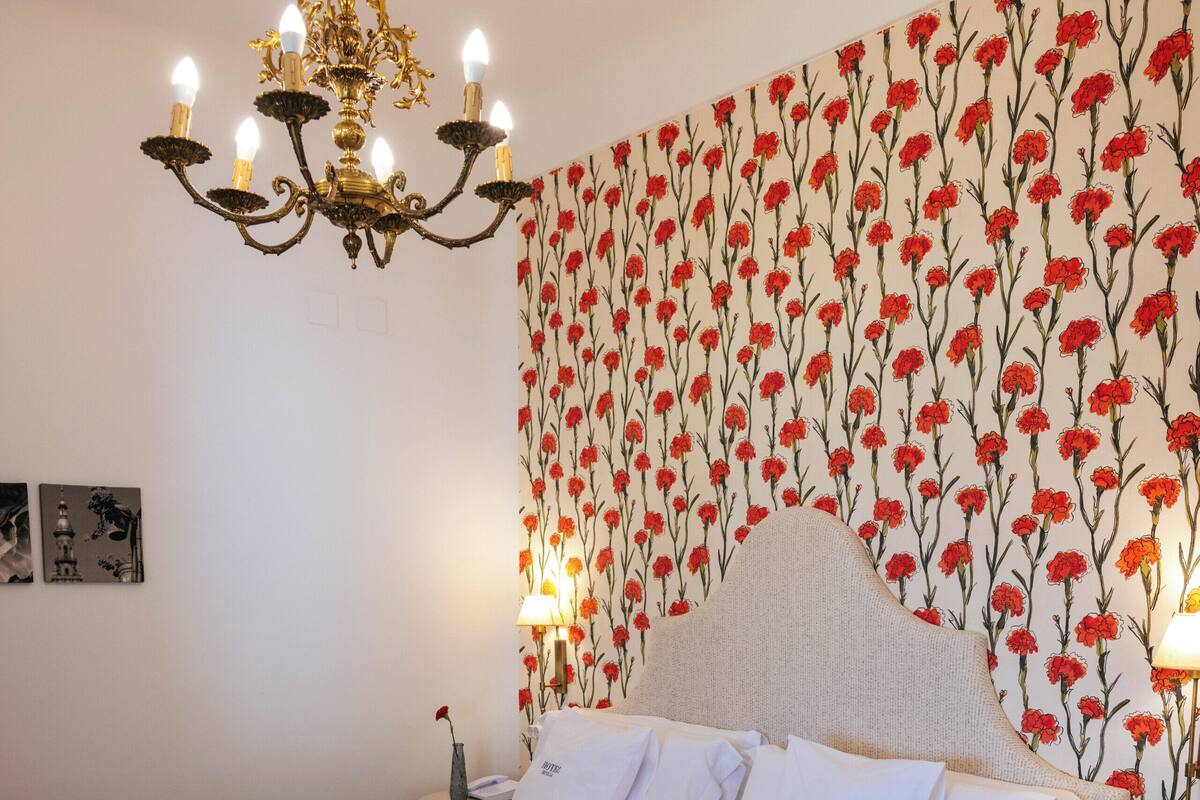 Ein Doppelbett mit Kopfteil, eine Chandelüster, ein Nachttisch und eine Wand mit floralem Wandpapier.