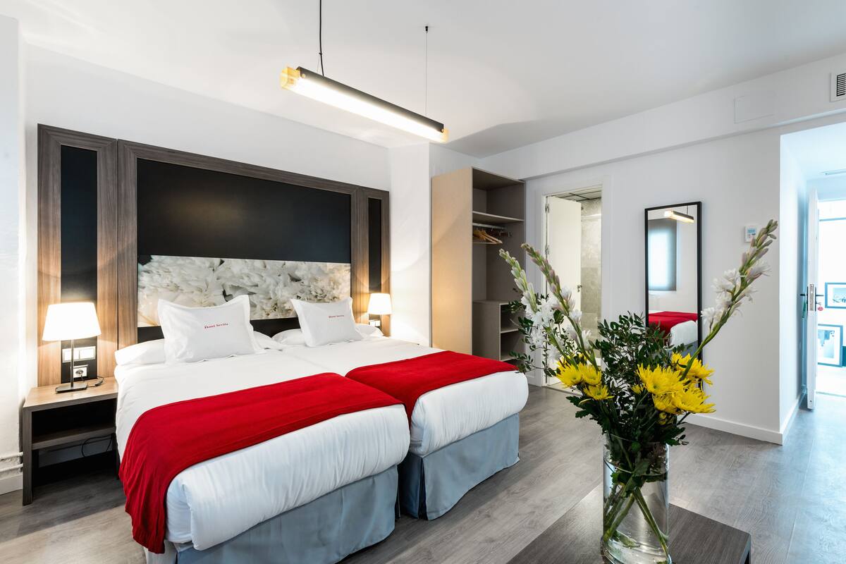 Ein modernes Hotelzimmer mit einem großen Bett, einem Nachttisch mit Lampe, einem Spiegel und einer Vase mit Blumen.