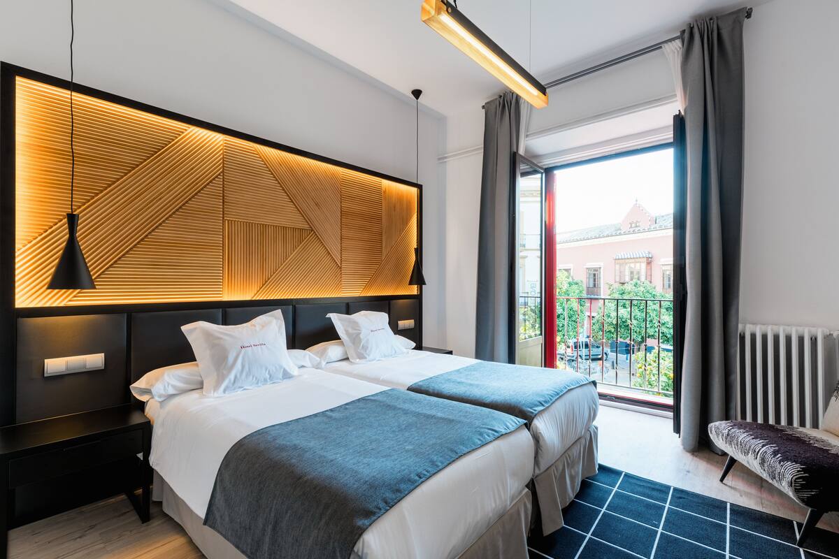 Ein modernes Hotelzimmer mit zwei Betten, einem Kopfteil mit geometrischem Muster, Blick auf die Stadt und einem Balkon mit Blick auf Gebäude.