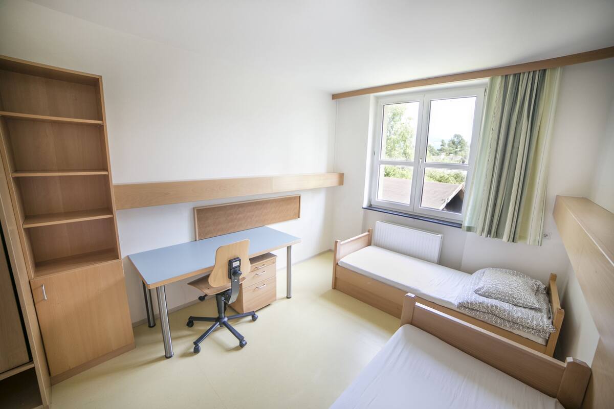 A bedroom with a wooden desk, a bed, a window with curtains, and a built-in shelf.