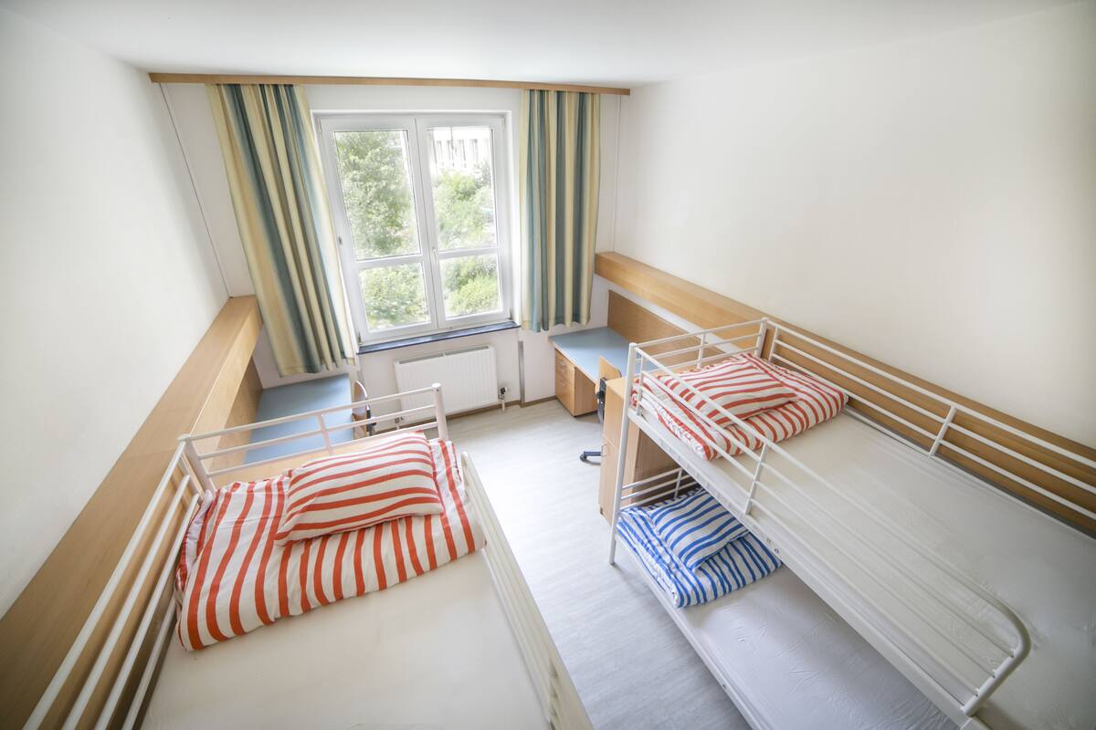 A dormitory room with bunk beds, a window with curtains, and a desk.