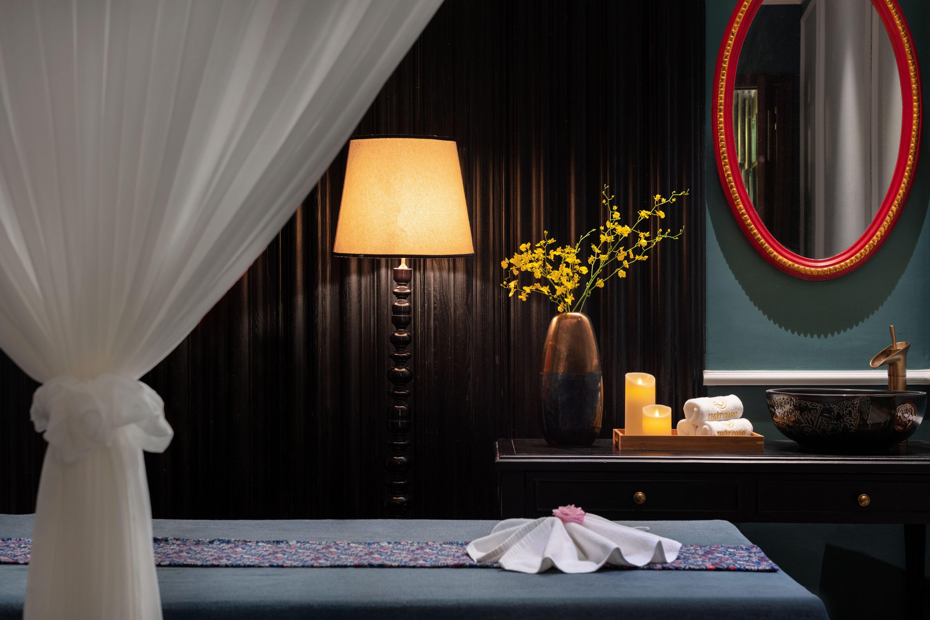 A bedroom with a bed, a bedside table with a lamp, a vase with flowers, candles, and a mirror.