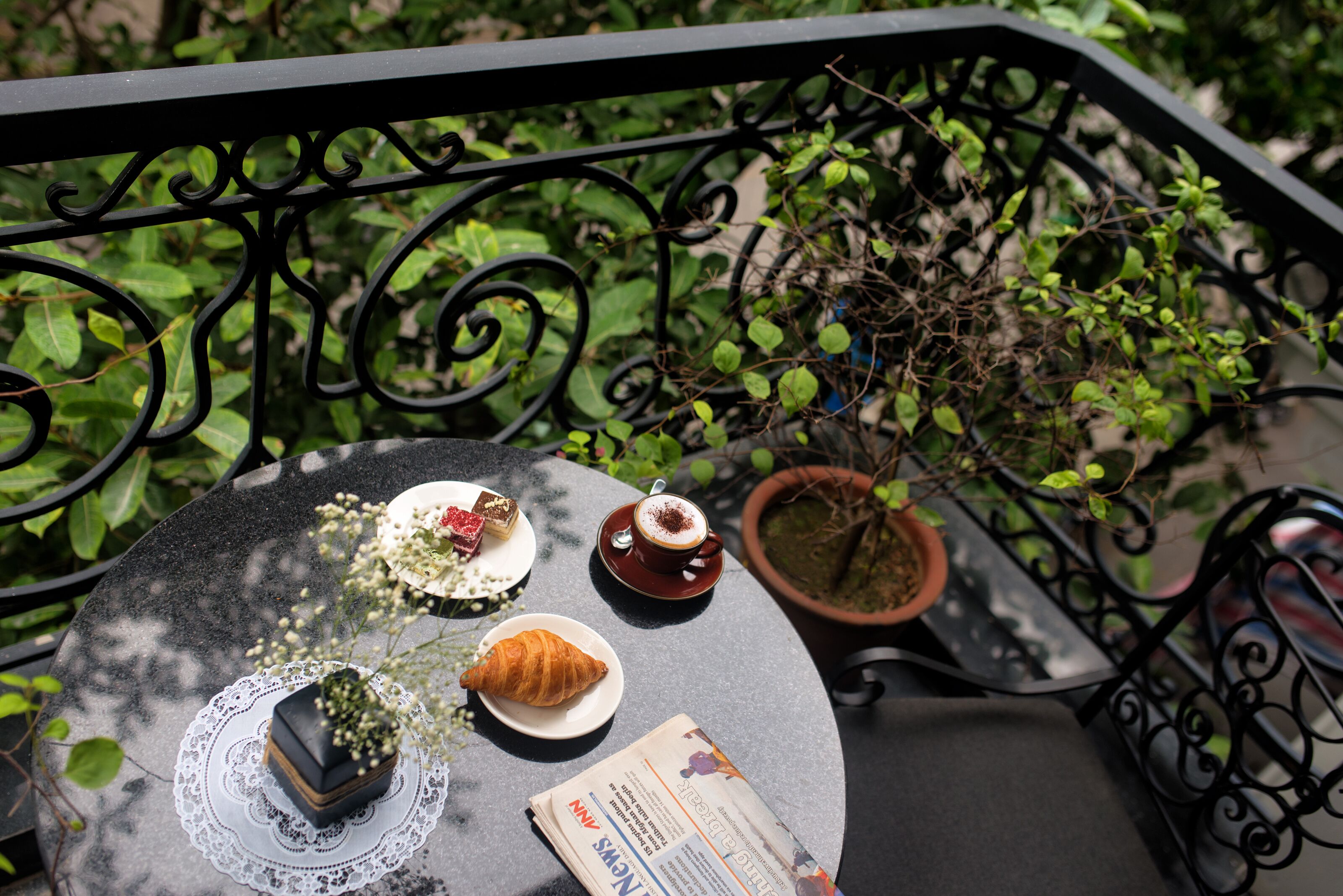 A round table with a croissant, a dessert, and a cup of coffee on a terrace with a potted plant and a railing.