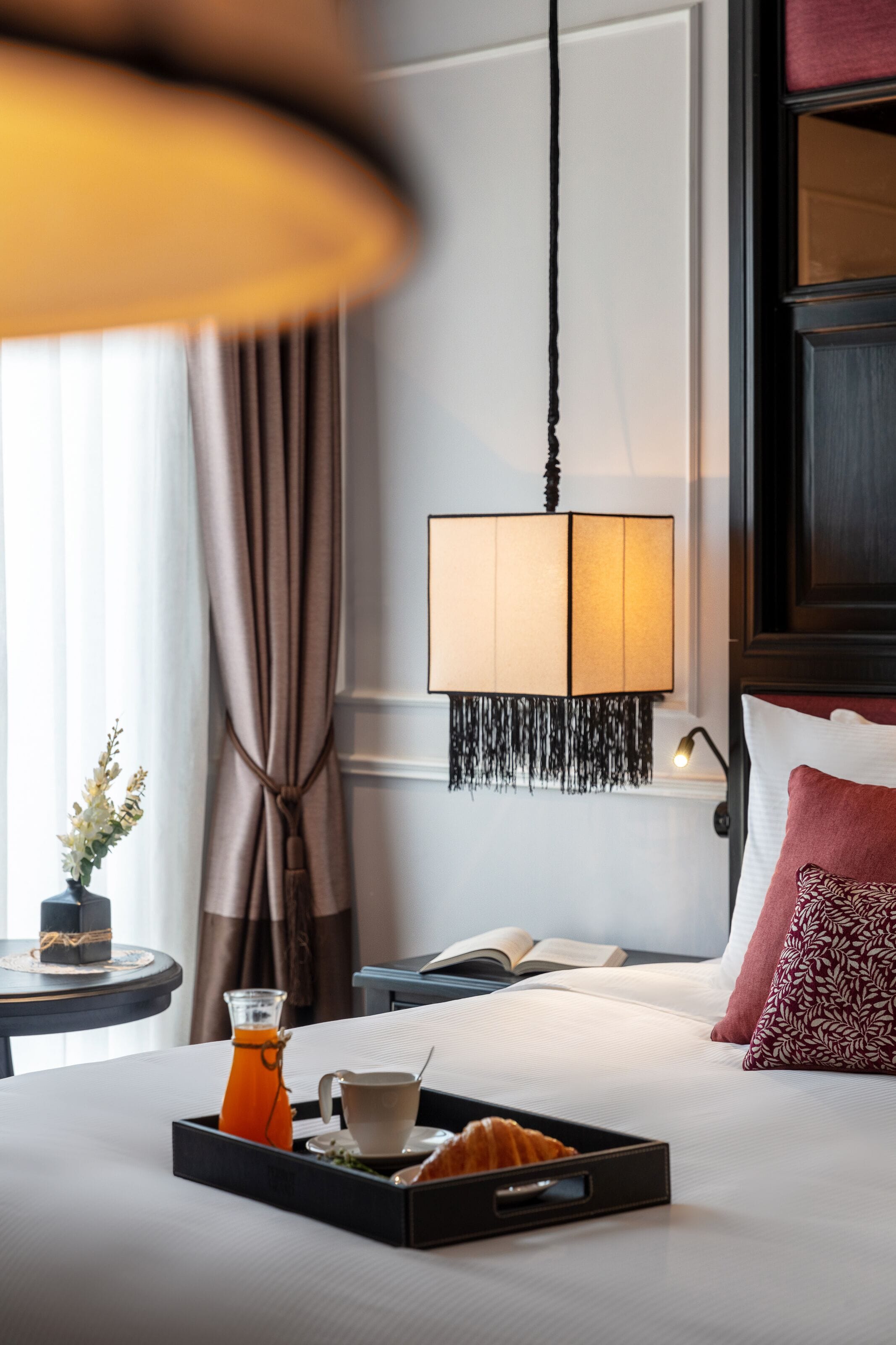 Une chambre d’hôtel avec un lit, une table de chevet sur laquelle se trouve un plateau de petit-déjeuner, une lampe et un livre.