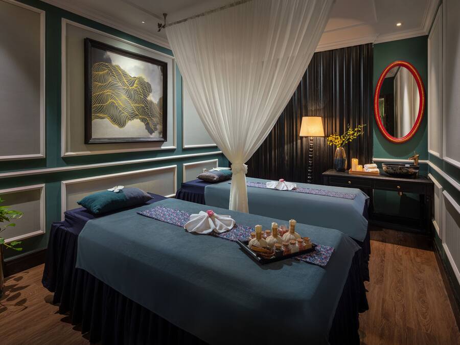Une chambre d’hôtel avec deux lits, un baldaquin, un tableau encadré, un miroir et une table de chevet avec une lampe et des bougies.