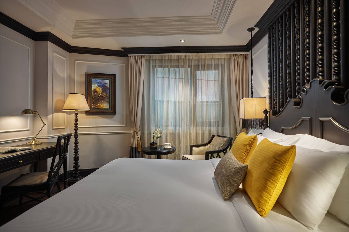 A hotel room with a large bed, two bedside tables, a desk, a chair, and a window with sheer curtains.