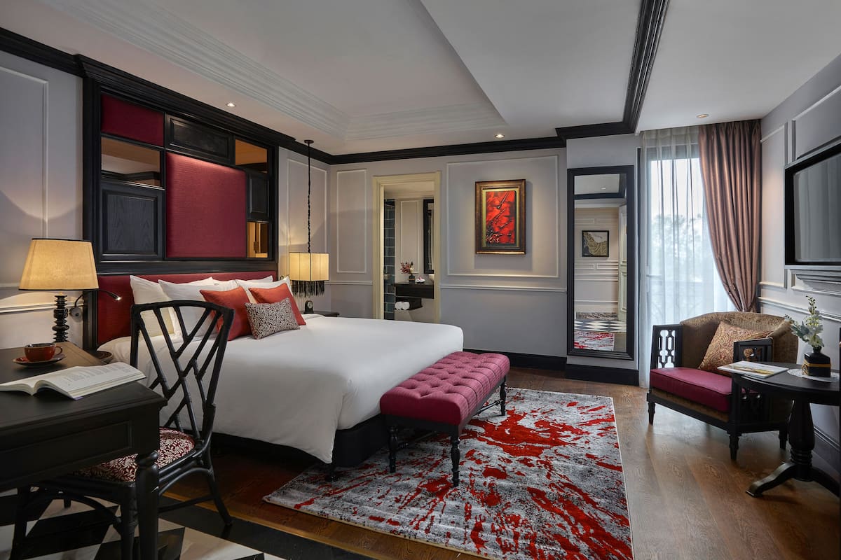 A hotel room with a large bed, a desk with a lamp, a chair, a red rug, and a sofa.