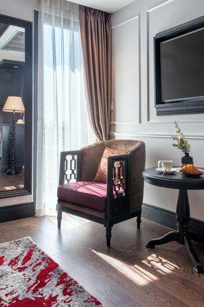 A modern hotel room with a dark wood floor, a red and white patterned rug, a dark wood armchair with a red cushion, a small round table with a white cup and a croissant, and a large flat-screen TV mounted on the wall.