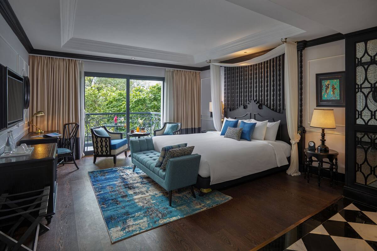 A hotel room with a large bed, a desk, a chair, a small table, and a balcony with a view of greenery.
