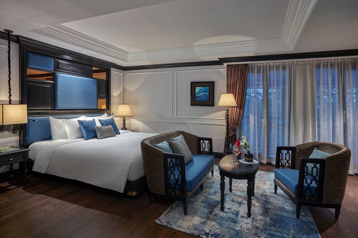 A hotel room with a large bed, two armchairs, a small table, and a large window with curtains.