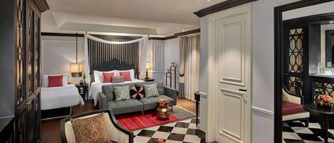 Una habitación de hotel con piso a cuadros, una cama con ropa de cama blanca y almohadas rojas, un sofá gris con cojines estampados, un sillón rojo y una pequeña mesita de noche con un florero.