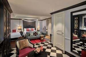 A hotel room with a checkered floor, a bed with white bedding and red pillows, a grey sofa with patterned cushions, a red armchair, and a small side table with a vase.