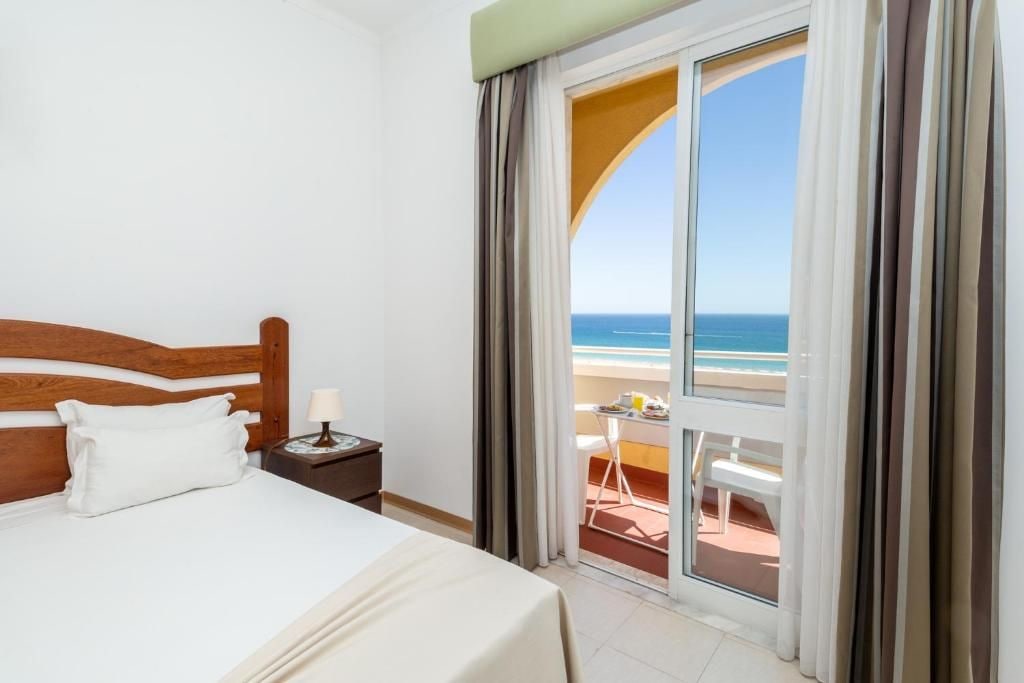A hotel room with a bed, a nightstand, a lamp, and a view of the beach through a large window.