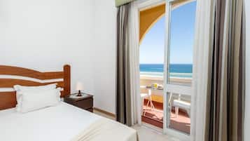 A hotel room with a bed, a nightstand, a lamp, and a view of the beach through a large window.