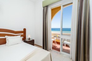 Habitación de hotel con cama, mesita de noche, lámpara y vista a la playa a través de un ventanal grande.