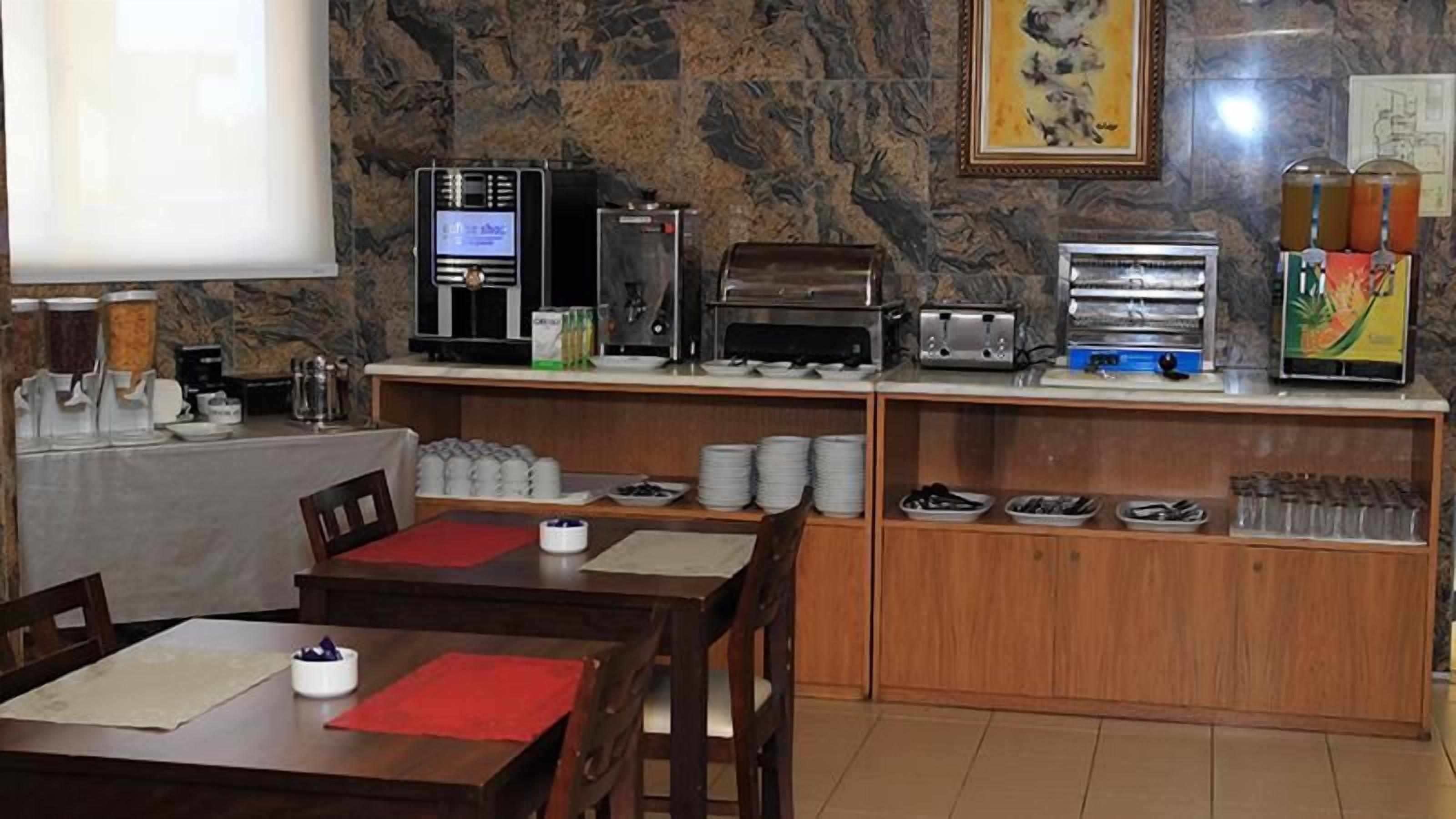 A breakfast area with a coffee machine, toaster, and a beverage dispenser. There are tables with red placemats and chairs.
