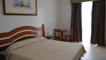 A hotel room with a large bed, a wooden headboard, a small table with a chair, a nightstand, and a window with curtains.